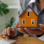 Casa laranja, segurada por uma pessoa com roupa formal; entenda as regras do financiamento imobiliário Casa laranja, segurada por uma pessoa com roupa formal; entenda as regras do financiamento imobiliário