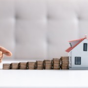 Mão segura moedas e as coloca em cima de outras, empilhadas em direção a uma casinha. Imagem remete ao financiamento imobiliário ou a poupança de dinheiro para compra da casa própria. Mão segura moedas e as coloca em cima de outras, empilhadas em direção a uma casinha. Imagem remete ao financiamento imobiliário ou a poupança de dinheiro para compra da casa própria.
