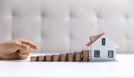 Mão segura moedas e as coloca em cima de outras, empilhadas em direção a uma casinha. Imagem remete ao financiamento imobiliário ou a poupança de dinheiro para compra da casa própria. Mão segura moedas e as coloca em cima de outras, empilhadas em direção a uma casinha. Imagem remete ao financiamento imobiliário ou a poupança de dinheiro para compra da casa própria.
