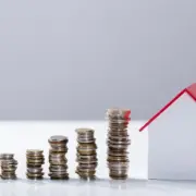 Imagem mostra pilha de moedas crescendo em direção a um cofre em formato de casa, que está sendo abastecido com mais uma moeda por uma pessoa. Remete à ideia de financiamento imobiliário, como o SFH. Imagem mostra pilha de moedas crescendo em direção a um cofre em formato de casa, que está sendo abastecido com mais uma moeda por uma pessoa. Remete à ideia de financiamento imobiliário, como o SFH.