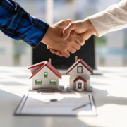 Aperto de mãos entre corretor e cliente sobre maquetes de casas e contrato de compra, simbolizando tecnologia e liderança no mercado imobiliário Aperto de mãos entre corretor e cliente sobre maquetes de casas e contrato de compra, simbolizando tecnologia e liderança no mercado imobiliário