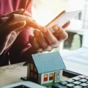 Pessoa contrata algo em um celular. Diante dela, uma calculadora e uma casinha, remetendo à ideia de contrato relacionado ao mercado imobiliário. Pessoa contrata algo em um celular. Diante dela, uma calculadora e uma casinha, remetendo à ideia de contrato relacionado ao mercado imobiliário.