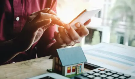 Pessoa contrata algo em um celular. Diante dela, uma calculadora e uma casinha, remetendo à ideia de contrato relacionado ao mercado imobiliário. Pessoa contrata algo em um celular. Diante dela, uma calculadora e uma casinha, remetendo à ideia de contrato relacionado ao mercado imobiliário.