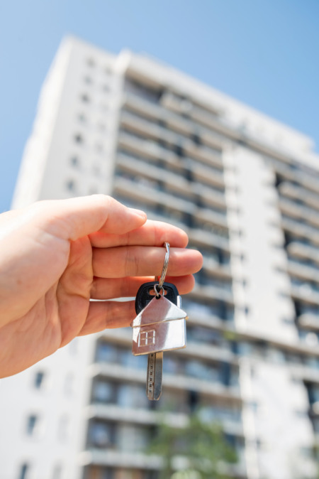 Mão segurando chave com chaveiro em formato de casa em frente a prédio residencial Mão segurando chave com chaveiro em formato de casa em frente a prédio residencial