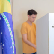 Bandeira do Brasil em primeiro plano e eleitor votando em cabina da Justiça Eleitoral ao fundo Bandeira do Brasil em primeiro plano e eleitor votando em cabina da Justiça Eleitoral ao fundo