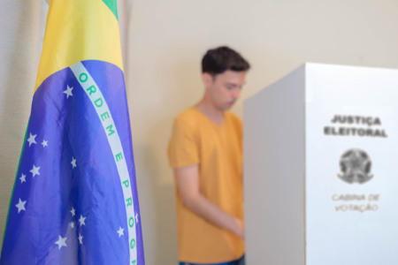 Bandeira do Brasil em primeiro plano e eleitor votando em cabina da Justiça Eleitoral ao fundo Bandeira do Brasil em primeiro plano e eleitor votando em cabina da Justiça Eleitoral ao fundo