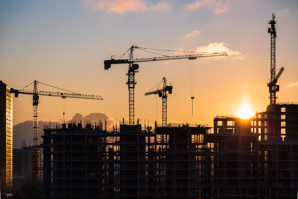 Guindastes em obra de edifício residencial ao pôr do sol, simbolizando a construção civil e o mercado imobiliário Guindastes em obra de edifício residencial ao pôr do sol, simbolizando a construção civil e o mercado imobiliário