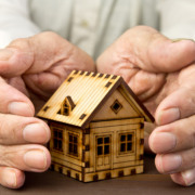 Mãos de um idoso protegendo uma maquete de casa de madeira, simbolizando moradia para a terceira idade Mãos de um idoso protegendo uma maquete de casa de madeira, simbolizando moradia para a terceira idade