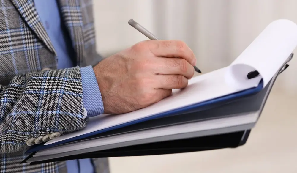 Mão segurando prancheta e fazendo apontamentos no papel, remetendo à ideia de vistoria técnica para elaboração de um laudo ou parecer, como o PTAM. Mão segurando prancheta e fazendo apontamentos no papel, remetendo à ideia de vistoria técnica para elaboração de um laudo ou parecer, como o PTAM.