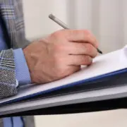 Mão segurando prancheta e fazendo apontamentos no papel, remetendo à ideia de vistoria técnica para elaboração de um laudo ou parecer, como o PTAM. Mão segurando prancheta e fazendo apontamentos no papel, remetendo à ideia de vistoria técnica para elaboração de um laudo ou parecer, como o PTAM.