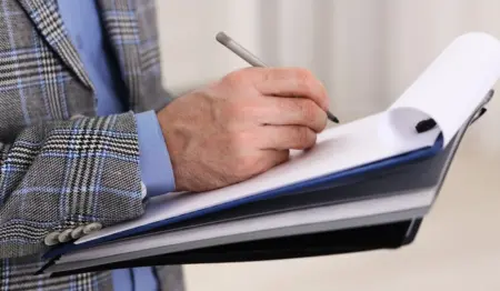 Mão segurando prancheta e fazendo apontamentos no papel, remetendo à ideia de vistoria técnica para elaboração de um laudo ou parecer, como o PTAM. Mão segurando prancheta e fazendo apontamentos no papel, remetendo à ideia de vistoria técnica para elaboração de um laudo ou parecer, como o PTAM.