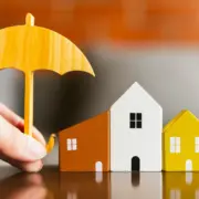 Guarda-chuva sendo segurado em cima de casinhas em miniatura, remetendo à ideia de seguro para garantir pagamento de financiamento imobiliário, como o seguro prestamista. Guarda-chuva sendo segurado em cima de casinhas em miniatura, remetendo à ideia de seguro para garantir pagamento de financiamento imobiliário, como o seguro prestamista.