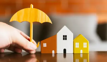 Guarda-chuva sendo segurado em cima de casinhas em miniatura, remetendo à ideia de seguro para garantir pagamento de financiamento imobiliário, como o seguro prestamista. Guarda-chuva sendo segurado em cima de casinhas em miniatura, remetendo à ideia de seguro para garantir pagamento de financiamento imobiliário, como o seguro prestamista.