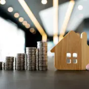 Monte de moedas empilhadas ao lado de uma casinha de madeira em miniatura, remetendo à ideia de financiamento imobiliário, como o SFI. Monte de moedas empilhadas ao lado de uma casinha de madeira em miniatura, remetendo à ideia de financiamento imobiliário, como o SFI.