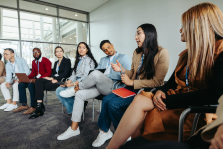 Grupo de profissionais reunidos em sessão de treinamento ou workshop corporativo, com uma mulher falando e os demais ouvindo com cadernos e laptops Grupo de profissionais reunidos em sessão de treinamento ou workshop corporativo, com uma mulher falando e os demais ouvindo com cadernos e laptops